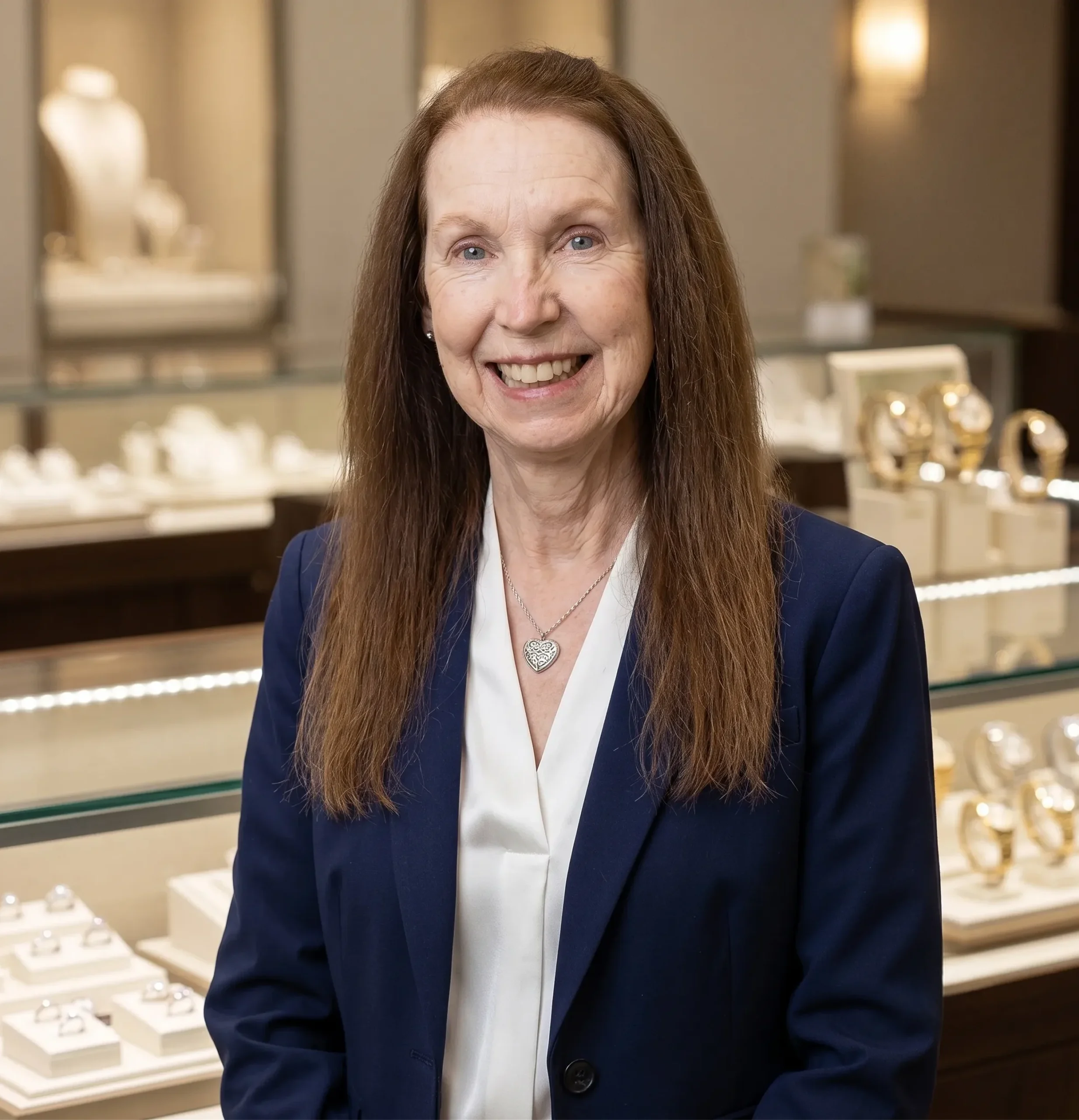 A woman with long brown hair and a warm smile, wearing a white blouse and a navy blue blazer, standing in front of a jewelry